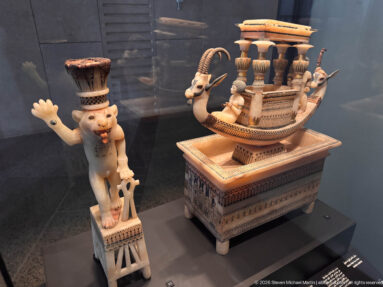 Display with an ornate boat-shaped polychrome object beside a standing dwarf-like figure wearing a tall headdress and raising one arm.