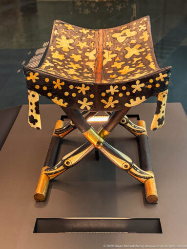 Ancient folding stool with X-shaped legs, dark concave seat, yellow floral motifs, and multicolored painted decoration.