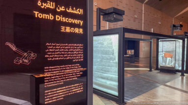 Multilingual “Discovery” panel beside cases containing stone architectural remains and an inscribed fragment in the museum gallery.