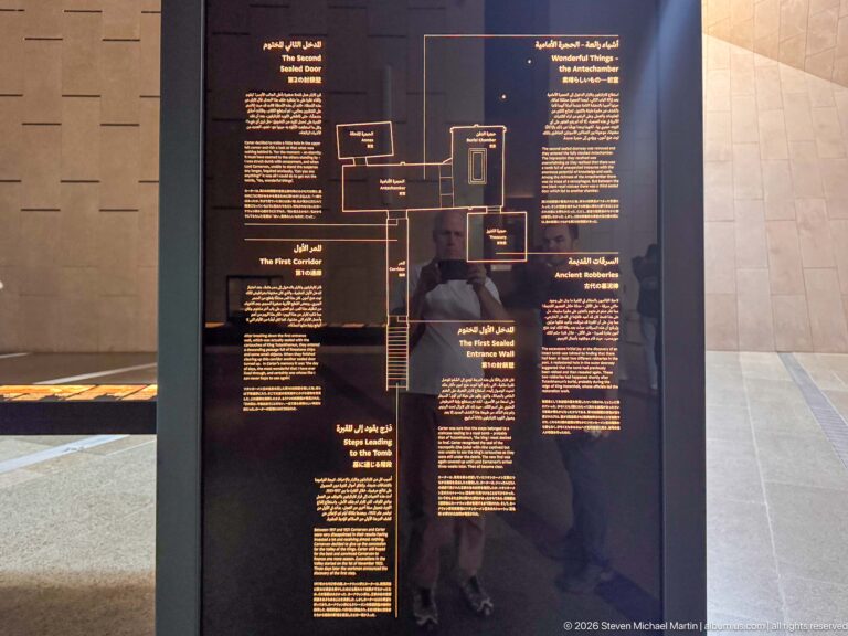 Black museum panel with a central diagram linking several Arabic and English text blocks about coffins and funerary preparation.