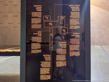 Black museum panel with a central diagram linking several Arabic and English text blocks about coffins and funerary preparation.