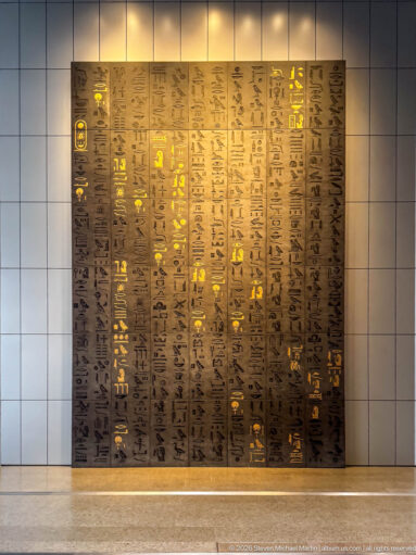Large backlit wall panel filled with vertical rows of Egyptian hieroglyphs, some glowing gold against a dark bronze-toned surface.