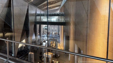 Elevated view into the Grand Egyptian Museum interior with tall columns, bridges, stone walls, and visitors far below.