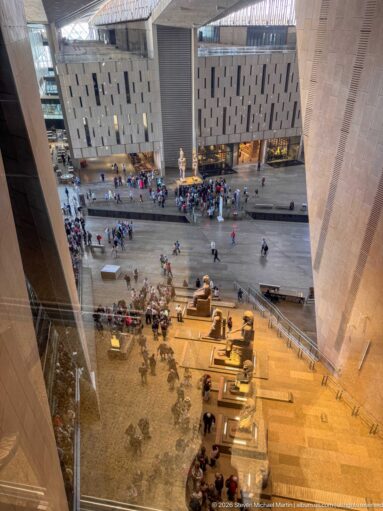 Elevated view into the Grand Egyptian Museum atrium with sloping stone walls, visitors, and monumental sculptures across the floor.