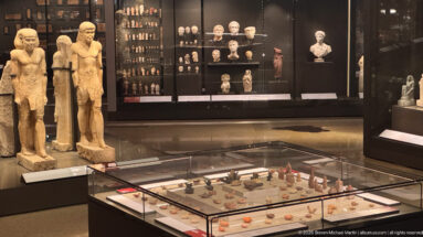 Gallery display with two tall standing statues, a wall case of sculpted heads, and a low vitrine of small artifacts.