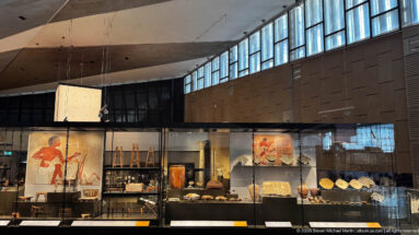 Wide Grand Egyptian Museum gallery view with a long glass case containing reliefs, vessels, sculpture, and small artifacts beneath high windows.