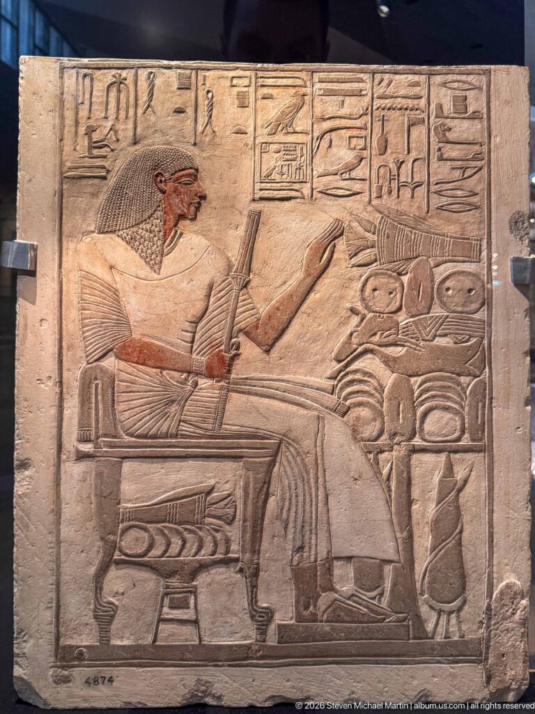 Painted limestone relief of a seated man facing provisions and vessels beneath hieroglyphic inscriptions.