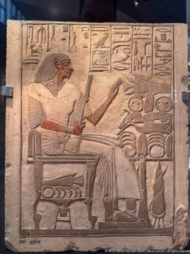 Painted limestone relief of a seated man facing provisions and vessels beneath hieroglyphic inscriptions.
