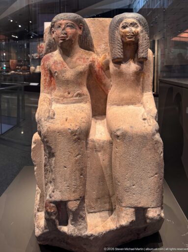 Stone seated couple on a shared bench, with dark wigs, traces of red pigment, and weathered surfaces.
