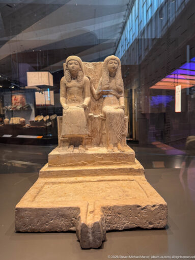 Stone group statue of two seated figures on a shared high-backed seat and rectangular base in the museum gallery.