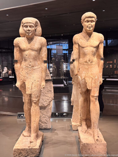 Two ancient Egyptian male statues standing side by side, both frontal with one leg advanced and visible surface losses.