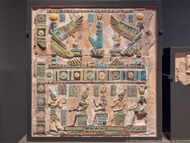 Painted Egyptian relief panel with two large winged figures above smaller ritual figures, hieroglyphs, and multicolored borders.
