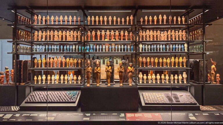 Wall-scale museum display with tightly ordered rows of shabtis in many colors, larger figures below, and small-object trays in front.
