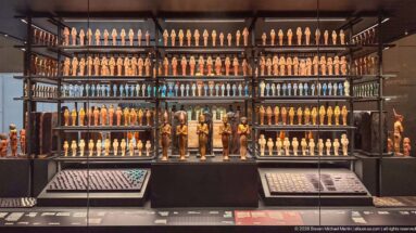 Wall-scale museum display with tightly ordered rows of shabtis in many colors, larger figures below, and small-object trays in front.