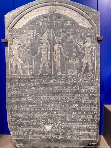 Dark rounded stone stela with carved figures above densely packed horizontal rows of hieroglyphic inscription.