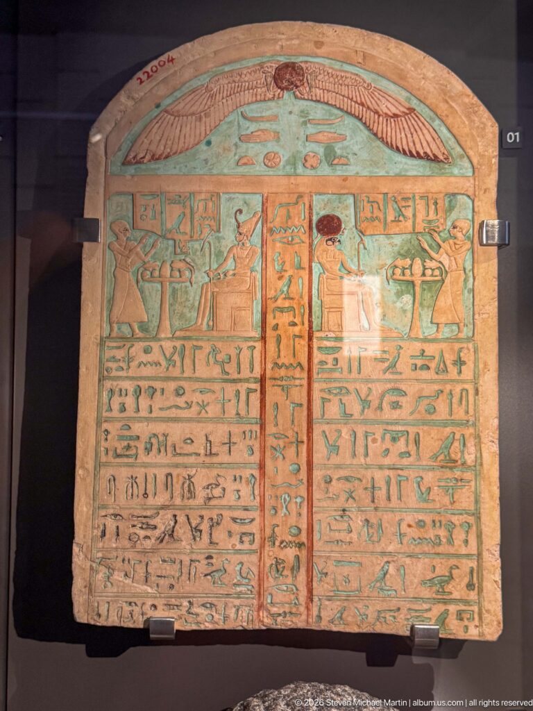 Rounded funerary stela with turquoise-blue ground, winged sun disk, hieroglyphic columns, and painted ritual figures.