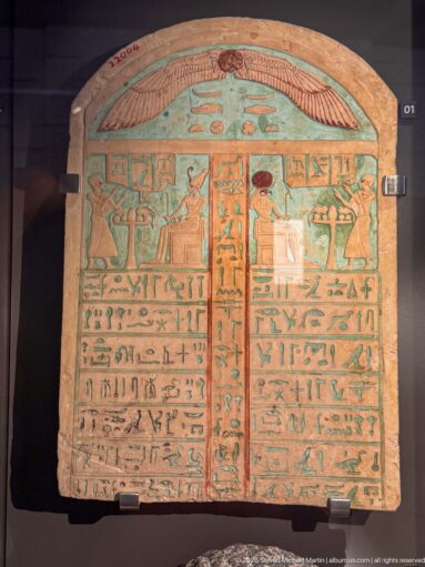 Rounded funerary stela with turquoise-blue ground, winged sun disk, hieroglyphic columns, and painted ritual figures.