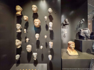 Gallery wall with many ancient sculpted heads on dark mounts and a larger painted female bust displayed in a case at right.