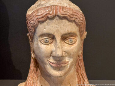 Close-up of a polychrome female sculpted head with wavy reddish hair, outlined eyes, softly modeled cheeks, and traces of surface color.