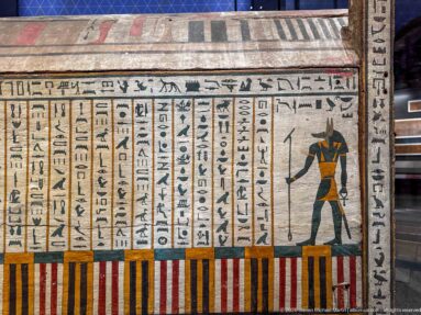 Painted funerary panel with vertical hieroglyphic columns, a standing jackal-headed deity, and striped lower border.