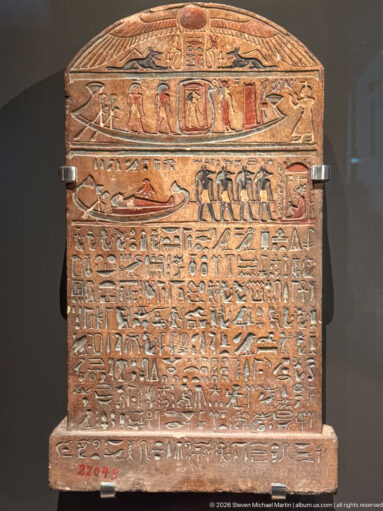 Rounded-top Egyptian funerary stela with painted figures, hieroglyphic registers, and dense inscription lines on an ochre surface.