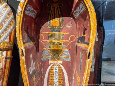 Painted coffin interior with central sacred emblem, ritual symbols, and hieroglyphic panels on a red ground.
