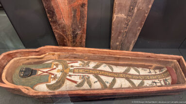 Wrapped ancient Egyptian mummy lying inside an open wooden anthropoid coffin, viewed from above.