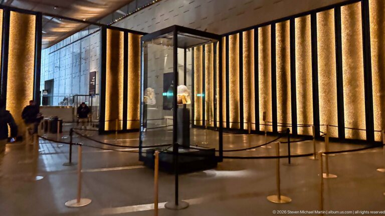 Wide Tutankhamun gallery with amber-lit wall panels, glass display case, rope barriers, and polished dark floor.