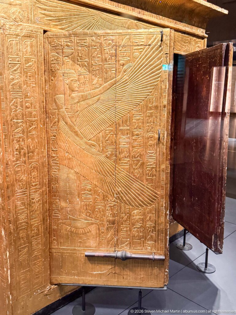 Open gilded shrine door engraved with a large winged goddess beside vertical hieroglyphs.