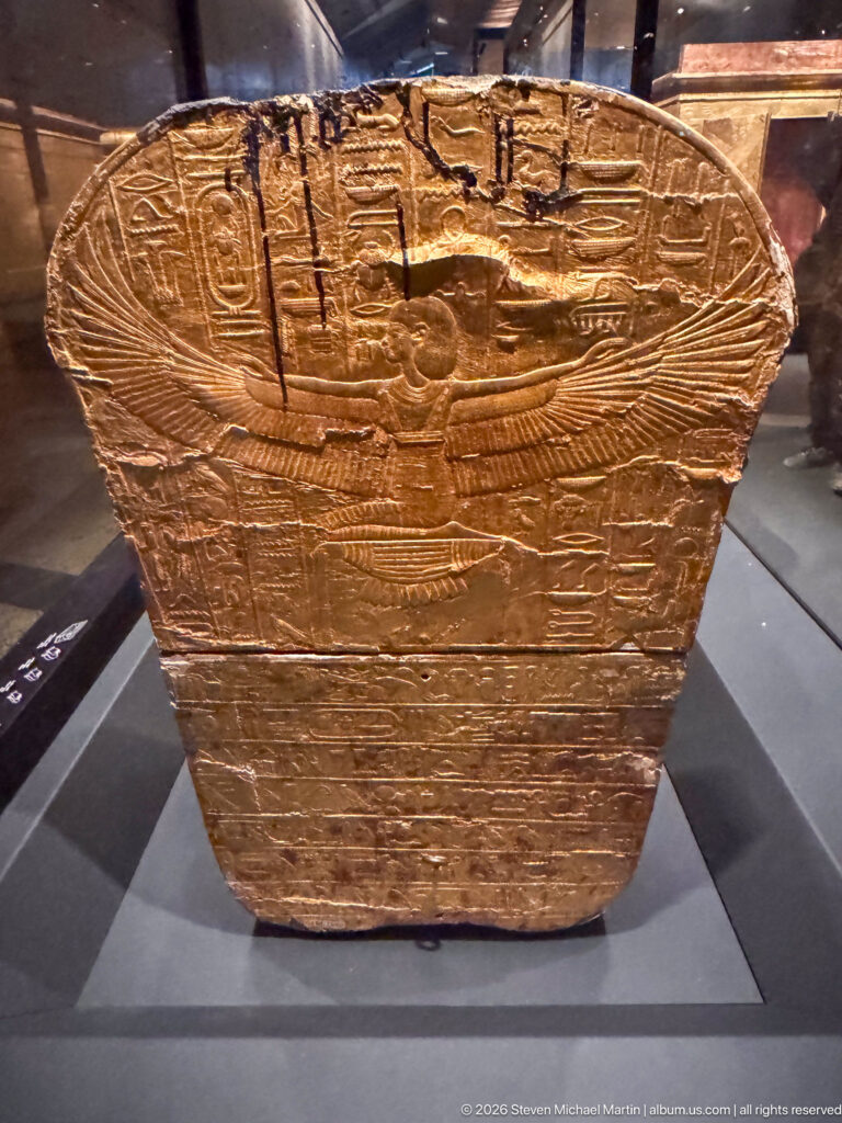 Rounded foot end of a gilded anthropoid coffin carved with feathers, wings, and horizontal bands.