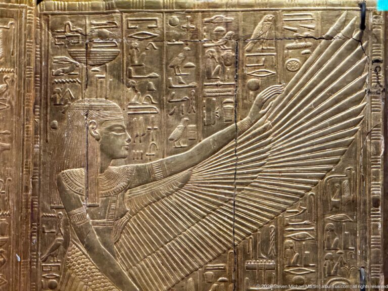 Winged Egyptian goddess extending a broad feathered wing across a gilded panel with hieroglyphs.