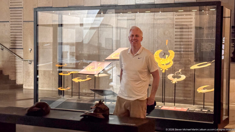 Visitor standing beside a glass case displaying large gold and multicolored broad collars.