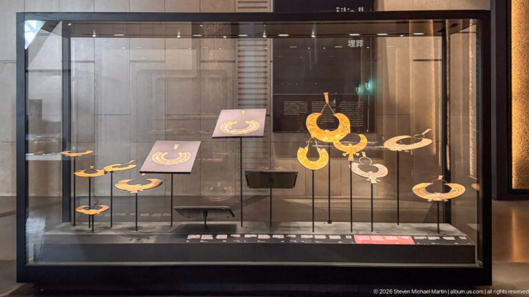 Display case with several large semicircular gold and multicolored broad collars mounted on stands.