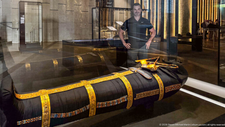 Side view of black wrapped anthropoid form with gold bands, colorful edging, and small chest ornaments.
