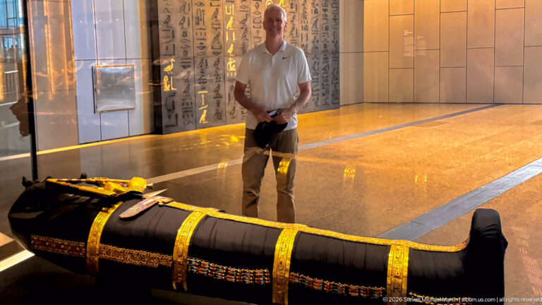 Black wrapped anthropoid body form with gold bands and chest ornaments, with a visitor standing behind the case.