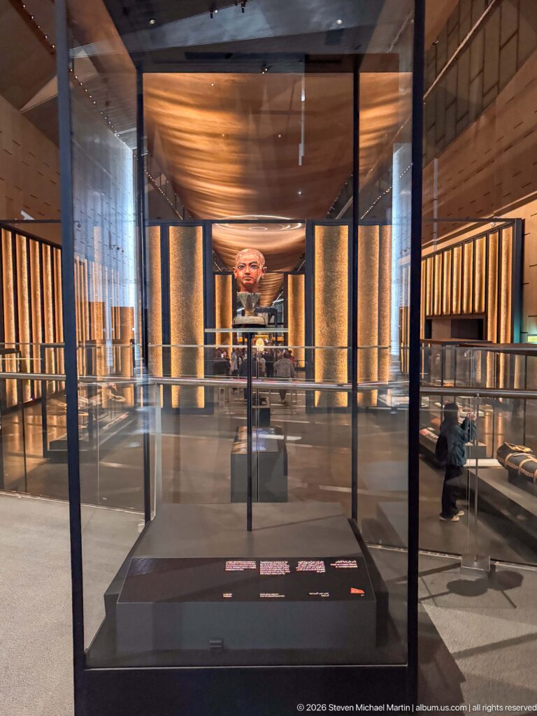 Wide gallery interior with glass case in foreground, illuminated columns, and Tutankhamun bust in the distance.
