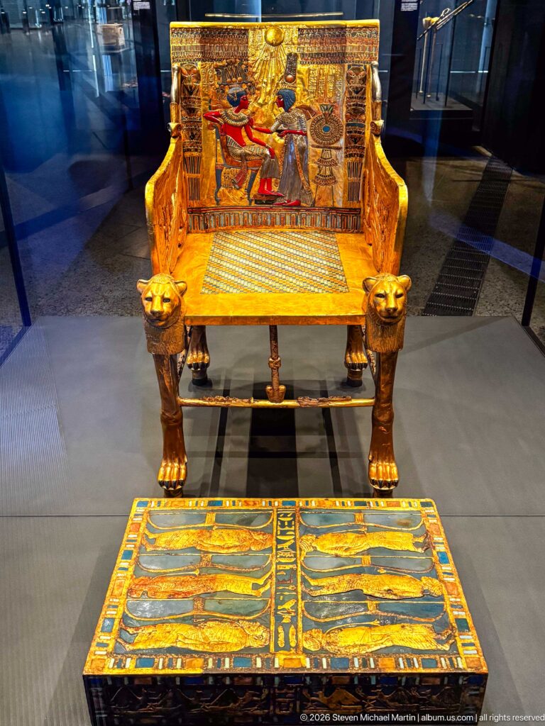 Front view of Tutankhamun’s Golden Throne with royal-couple backrest, gilded side panels, and decorated footstool.