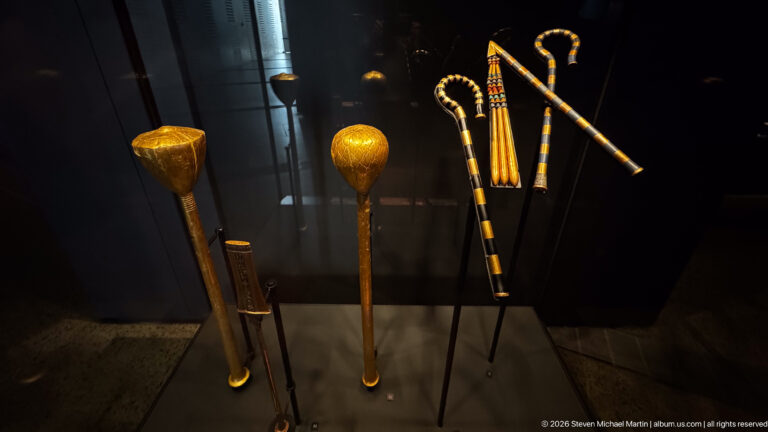 Long striped staffs, hooked crooks, and two bulbous ceremonial implements mounted against a dark background.