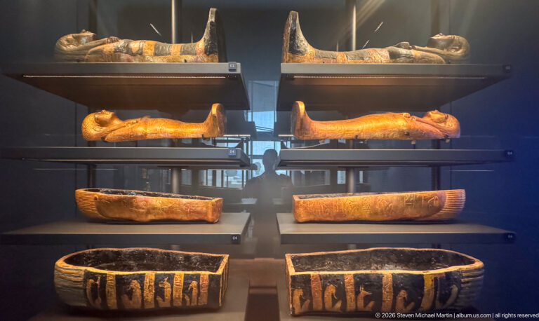 Stacked museum display with anthropoid coffins above and rectangular decorated funerary boxes below.