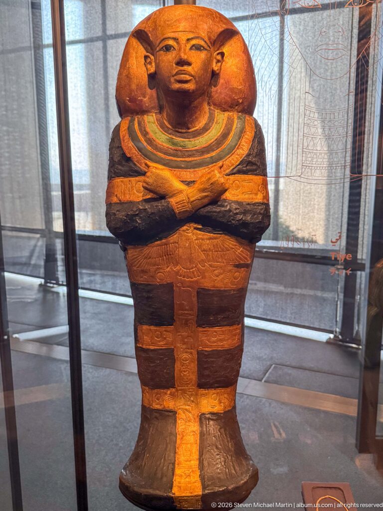 Large painted mummiform figure with crossed arms, broad multicolored collar, and dark-and-ochre body bands.