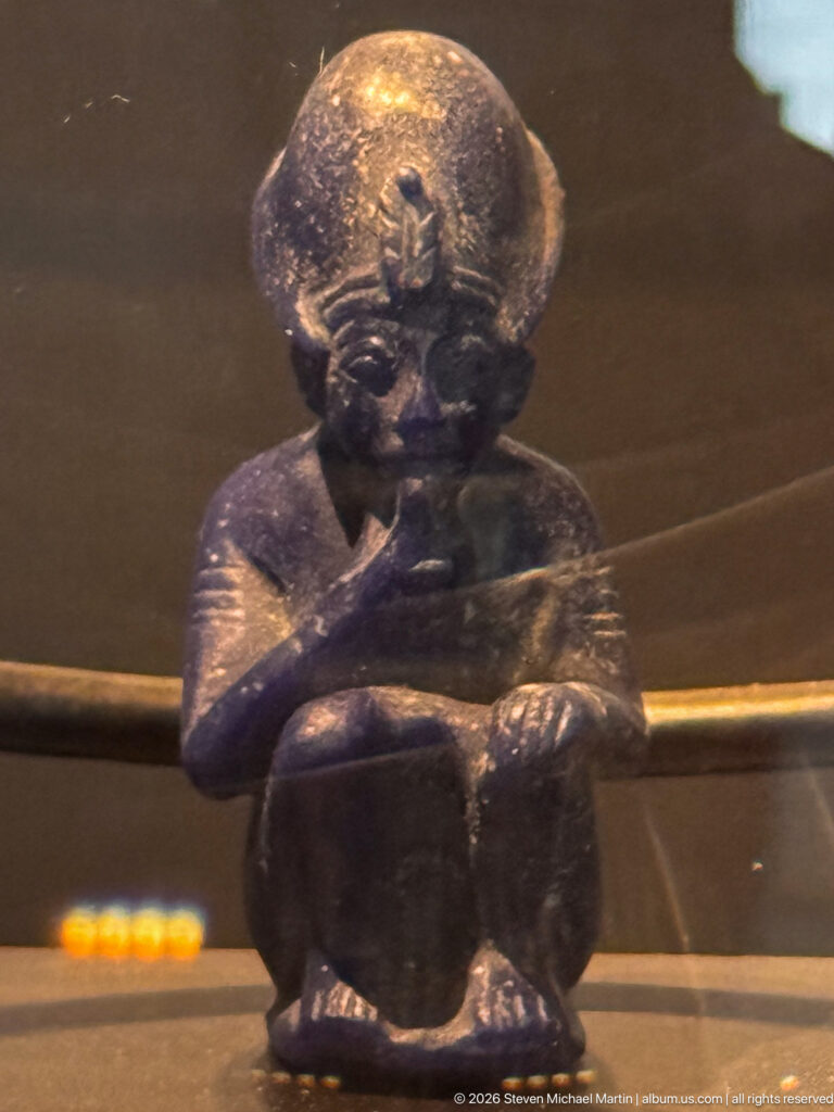 Small dark crouching or seated figure with rounded head and folded arms in a museum display.
