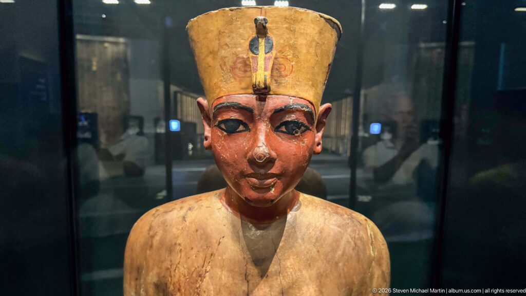 Painted wooden royal statue of Tutankhamun with tall crown, outlined eyes, and broad collar.