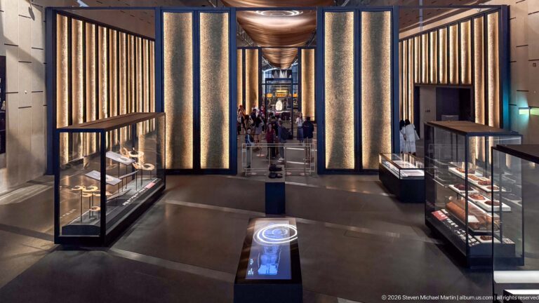 Long museum gallery with tall illuminated wall panels, glass display cases, and visitors in the distance.