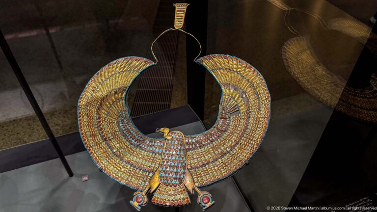 Oblique view of a multicolored winged falcon pectoral displayed behind reflective glass.