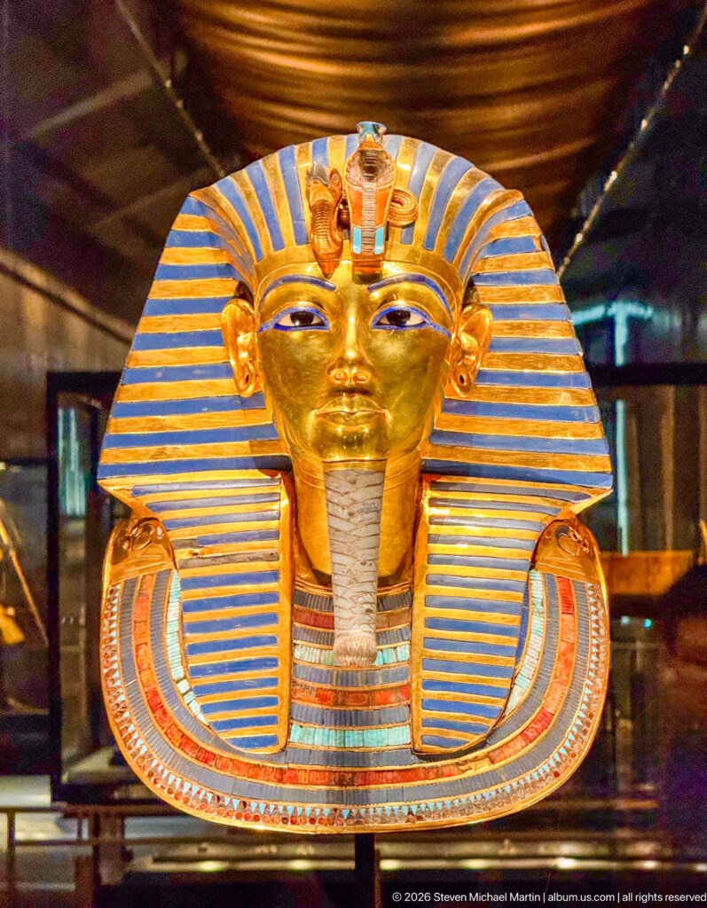 Frontal close view of Tutankhamun’s solid-gold burial mask with striped nemes, brow emblems, false beard, and inlaid collar.