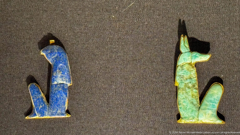 Blue seated animal amulet beside a turquoise-green jackal-form amulet on a dark display surface.