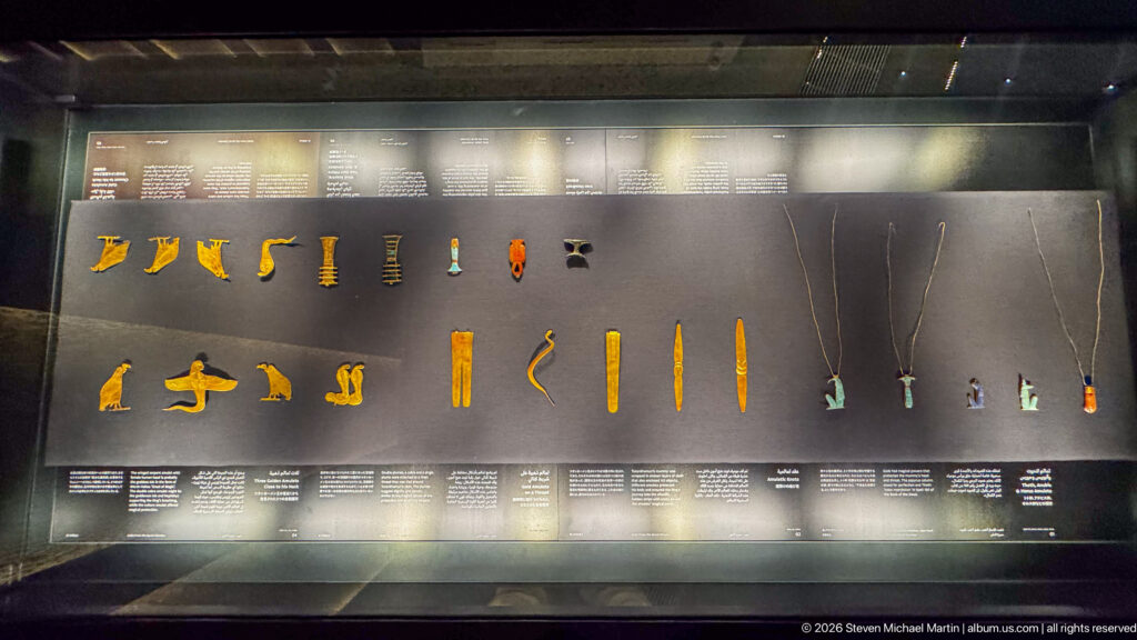Museum case filled with small gold amulets, colored pendants, and long necklaces arranged in rows.