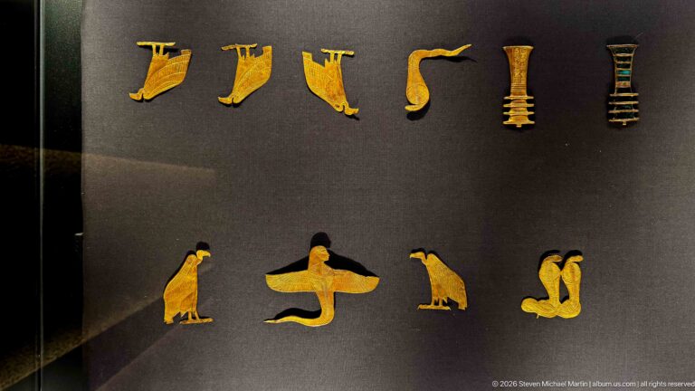 Rows of small gold and colored amulets in bird, serpent, and column-like forms on a dark display.