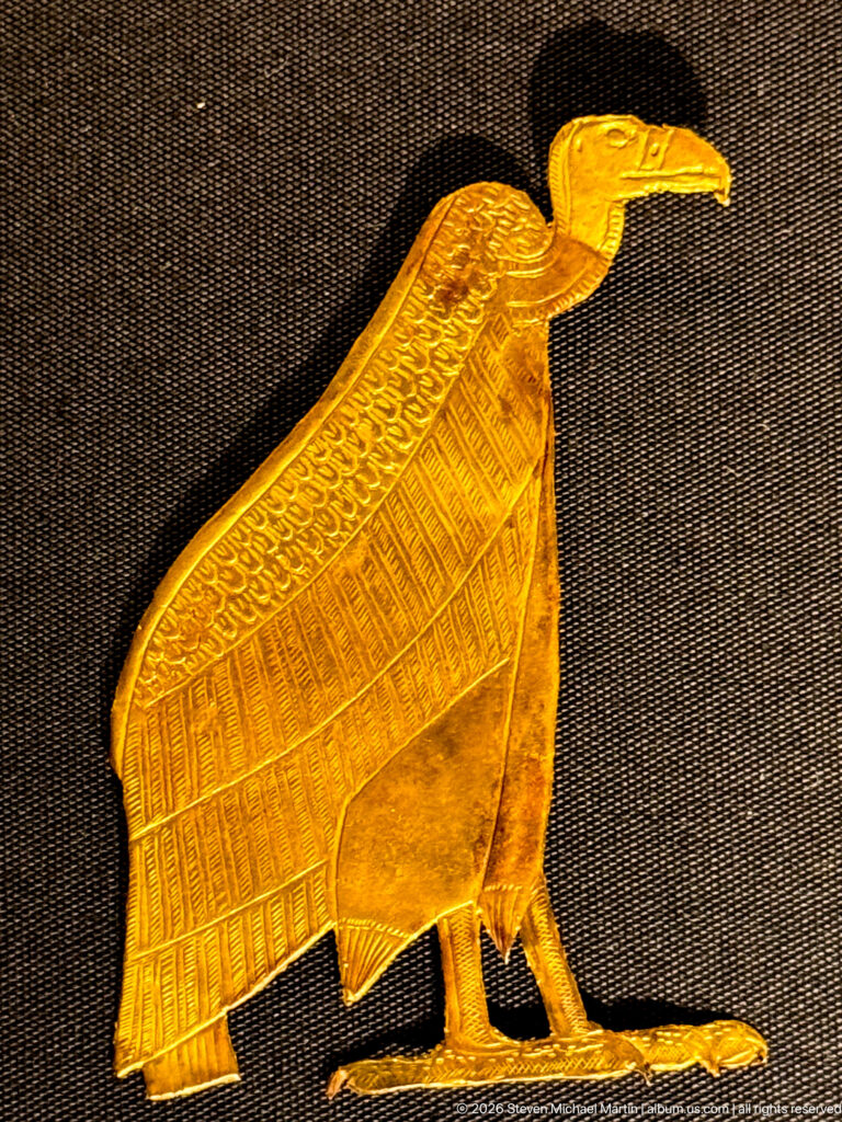 Standing gold vulture in profile with folded patterned wings and long projecting beak.