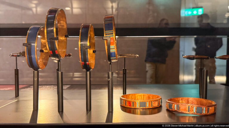 Several rigid gold bracelets and armlets with repeating blue and red inlays displayed on stands.
